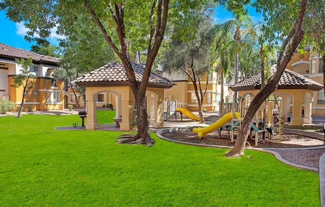 A playground with a yellow slide is surrounded by green grass and trees.