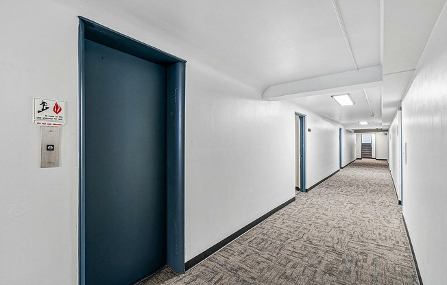 Modern apartment hallway with clean white walls, sleek blue elevator door, and bright overhead lighting.