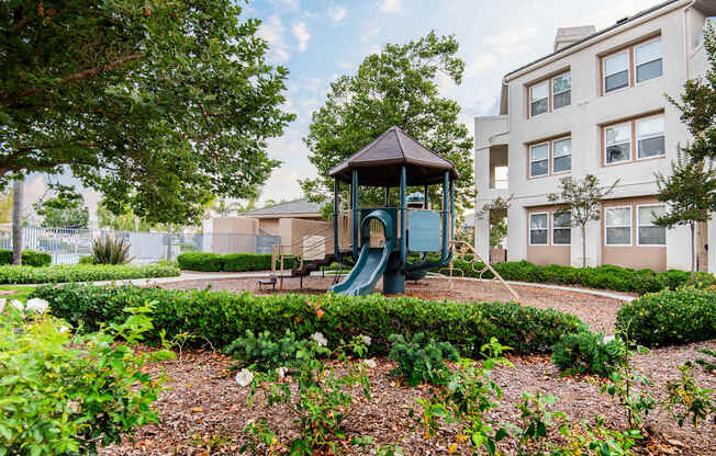 A playground with a green slide and a brown roof is surrounded by bushes and trees.