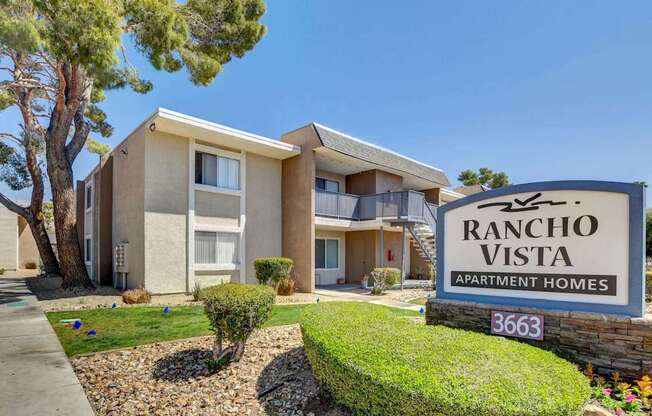 A Rancho Vista Apartment Homes sign is in front of a tan building with a tree and bushes in the front yard.