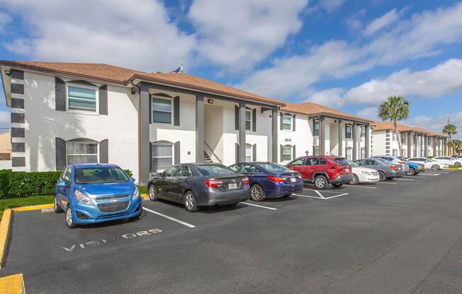 A well-maintained apartment complex featuring two-story white buildings with brown roofs. Multiple parked cars, including a blue hatchback and a red sedan, line the parking lot. Lush green landscaping is visible in front of the buildings under a partly cloudy sky.
