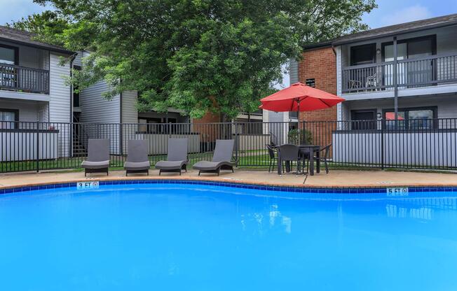 A clear blue swimming pool surrounded by a grassy area, featuring several lounge chairs and a table with an umbrella. In the background, there are two-story residential buildings with balconies.