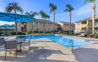 A sunny outdoor swimming pool area surrounded by palm trees, with lounge chairs and tables featuring blue umbrellas. The pool is clear and inviting, with residential buildings in the background, creating a relaxed atmosphere for leisure and recreation.