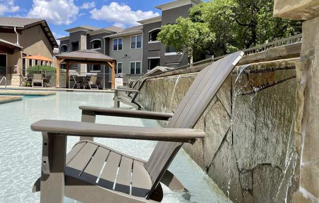 a lounge chair sits on the edge of a pool with a waterfall and buildings in the background