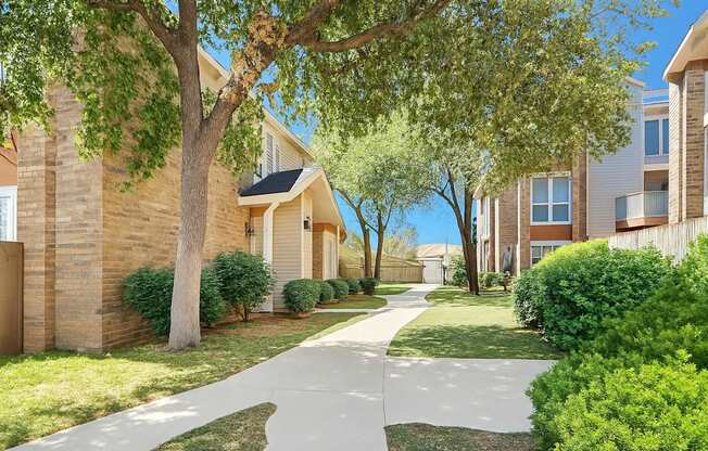 A peaceful landscaped walkway here at Summertree Place featuring a gently curving concrete path, mature shade trees, trimmed green shrubs, and well-maintained lawns surrounded by brick and siding apartment buildings, creating a quiet and inviting outdoor setting for residents to enjoy.