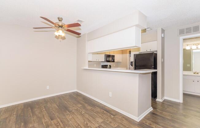 A modern living area featuring a ceiling fan, light-colored walls, and hardwood-style flooring. There is a breakfast bar with a view into a kitchen equipped with black appliances. The layout is open and spacious, creating a bright and inviting atmosphere.