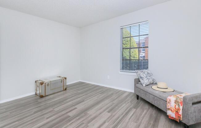 A spacious, empty room featuring light gray walls and a large window with blinds. There is a stylish gray couch with a decorative pillow and a sun hat on it. Next to the couch, there is a vintage suitcase, adding a touch of character to the minimalist space. The flooring is light-colored laminate.