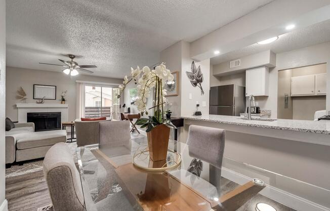 A modern living space featuring a dining area with a glass table and grey upholstered chairs, a cozy seating area with a light sofa, and a gas fireplace. The kitchen is visible in the background, showcasing granite countertops and stainless steel appliances. Decorative elements include a potted orchid and wall art.