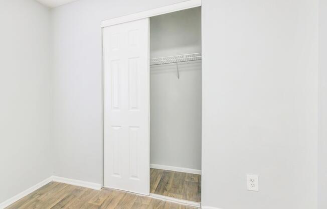 A small, empty room with light gray walls and a wooden floor. There is a closed sliding closet door on the left side, revealing a simple wire shelf inside. The overall atmosphere is clean and minimalist, with no decorations or furniture present.
