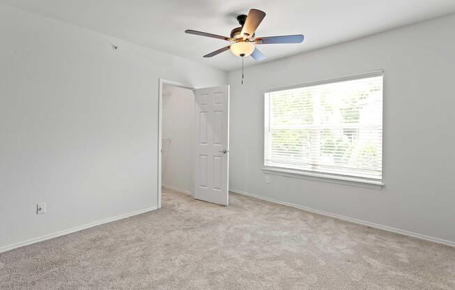 A spacious, empty room with light-colored walls and carpet. There is a ceiling fan with four blades and a light fixture. A window provides natural light, featuring white vertical blinds. An open door leads to a closet area, enhancing the sense of openness in the room.