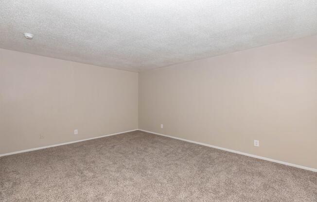 Empty room with beige walls and a light carpet. The ceiling is textured, and there are no furnishings or decorations. The space is well-lit, creating a clean and open atmosphere.