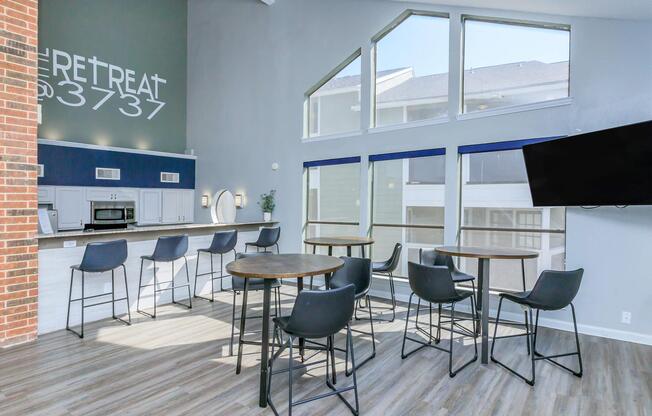 A modern lounge area featuring a bar with high stools, a kitchenette, and large windows allowing natural light. The walls are painted in light colors, and there is a television mounted on one wall. The space has a welcoming and contemporary atmosphere suitable for social gatherings.