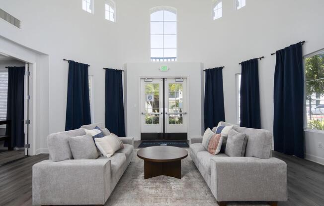 Bright, spacious living area featuring two gray sofas with colorful throw pillows, a round coffee table, and large windows allowing natural light. Blue curtains frame the windows, adding contrast to the white walls and wood flooring. A welcoming atmosphere with a modern design aesthetic.