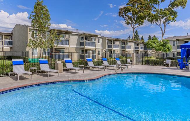 A sparkling blue swimming pool surrounded by lounge chairs under partly cloudy skies. In the background, there are apartment buildings with green landscaping and trees. The scene captures a relaxing outdoor space perfect for leisure and enjoyment.
