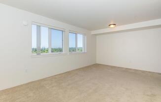 A spacious, empty room with light beige carpet and white walls. It features two large windows that let in natural light, offering a view of greenery outside. The ceiling is plain with a central light fixture. The room has a minimalist and bright atmosphere, suitable for various design possibilities.