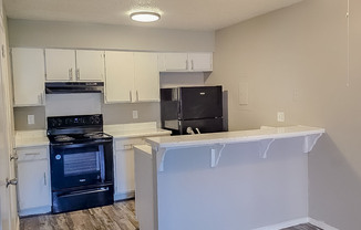 A modern kitchen featuring white cabinets, a black stove and refrigerator, and a bar counter with stools. The space is well-lit with a ceiling light and has a neutral wall color, creating an inviting atmosphere. The flooring is a light wood finish, enhancing the contemporary look.