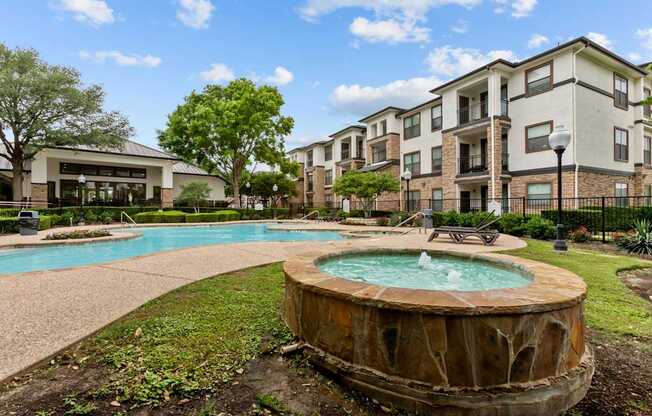 A large outdoor swimming pool with a hot tub in the middle of a grassy area in front of apartment buildings.
