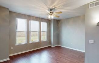 A bright empty living room here at BRIO at Firewheel with wood-style flooring, three tall windows with natural light, neutral walls, and a ceiling fan in an open, airy layout.