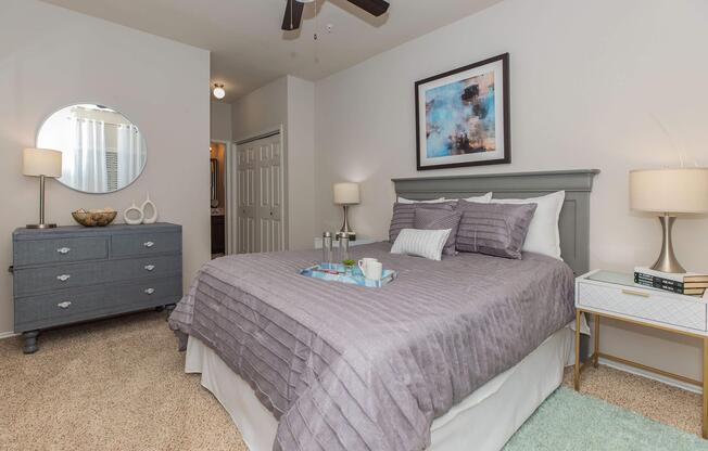 A neatly arranged bedroom featuring a gray upholstered bed with purple bedding, complemented by a gray dresser and a small nightstand. A round mirror hangs above the dresser, and there's soft lighting from the lamps. The room has light-colored walls and a cozy, inviting atmosphere.