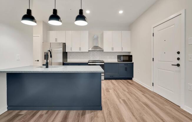 Modern kitchen featuring a central island with a blue base and light-colored countertops, stainless steel appliances, white upper cabinets, and dark blue lower cabinets. The room has three pendant lights hanging above the island, warm-toned wood flooring, and a doorway leading to an entrance.