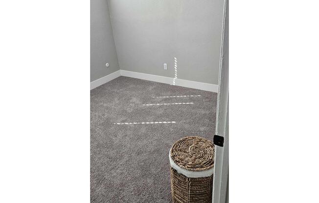 A sunlit corner of a room with light gray walls and plush carpet. A woven basket sits on the floor, and light streams in through a nearby door, casting a pattern on the carpet. The space appears empty and inviting, with a clean and minimalist aesthetic.