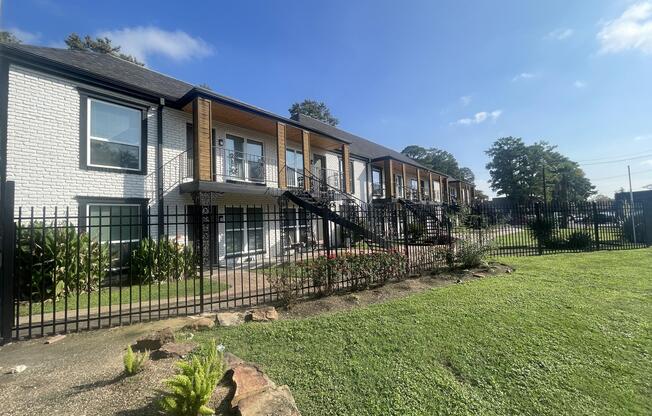 A row of modern apartment units with a mix of white brick and wooden accents. The buildings are surrounded by a black wrought-iron fence and feature stairs leading to upper levels. Lush green grass and shrubbery are present in the foreground under a clear blue sky.