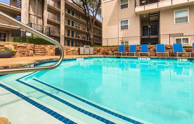 A pool area with lounge chairs, a patio table and chairs with a blue umbrella and stainless steel BBQ in front of apartment buildings at Princeton Court Apartments in the Vickery Midtown neighborhood of Dallas, TX.