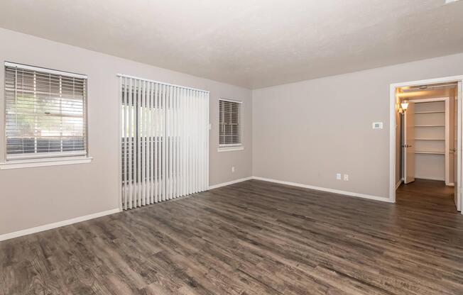 A spacious, empty room with gray walls and light wood flooring. Two windows with blinds on the left side allow natural light in. On the right, a doorway leads to a closet space. The overall atmosphere is bright and airy, suitable for various interior design options.