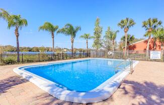 A clear blue swimming pool surrounded by palm trees and a fenced area. The sun shines brightly in a clear blue sky, with a serene lake visible in the background. The pool has a ladder for entry and is set on a patio made of tiles, creating a relaxing outdoor atmosphere.