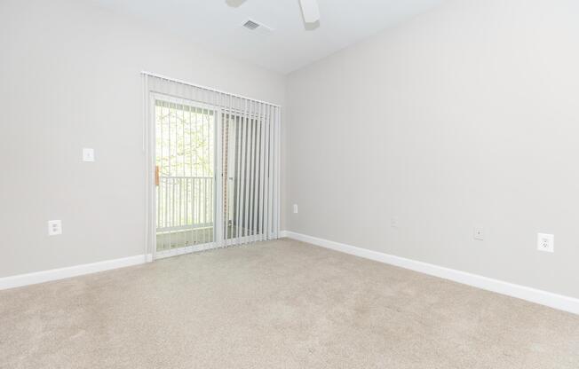 A spacious and empty room with light beige walls and a tan carpet. A sliding glass door with vertical blinds leads to a balcony, allowing natural light to enter. The room features minimal decor, emphasizing a clean and open layout.