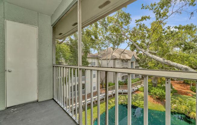 A balcony view overlooking a tranquil pond surrounded by greenery, with a building visible in the background. The railing and sliding door are visible, adding to the outdoor ambiance. Natural light filters through, creating a bright and inviting atmosphere.