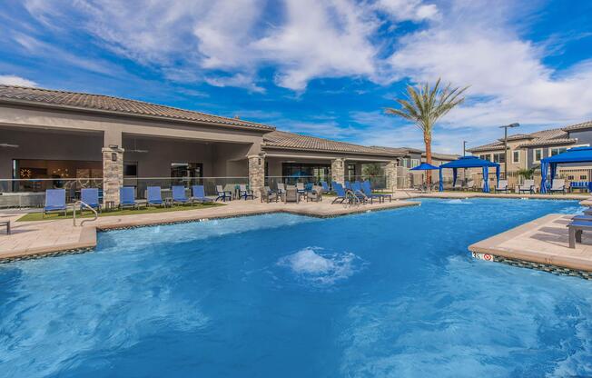 A sunny swimming pool area featuring a clear blue pool with a fountain, surrounded by lounge chairs and umbrellas. In the background, there are palm trees and a modern building with outdoor seating, all under a bright blue sky with scattered clouds.
