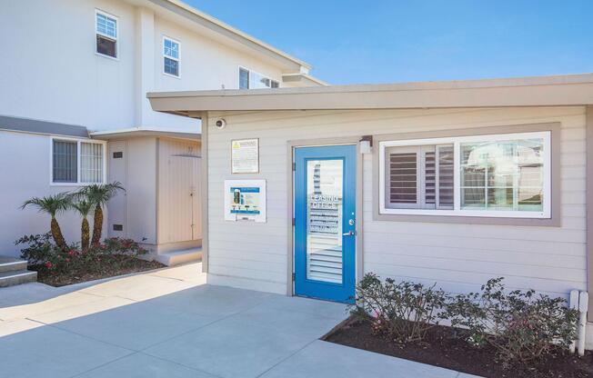 Exterior view of a modern building with a blue door, surrounded by shrubs and small palm trees. The entrance features a window with white shutters. In the background, another building is visible. The setting appears bright and sunny, highlighting the architectural design and landscaping.