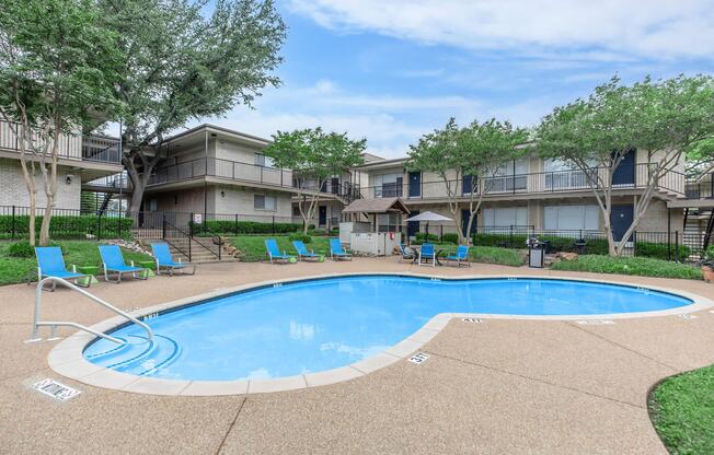 A well-maintained outdoor swimming pool surrounded by lounge chairs. The area features greenery with trees and two-story apartment buildings in the background. A shaded gazebo is visible near the poolside, creating a relaxing atmosphere. The sky is partly cloudy, enhancing the inviting setting.