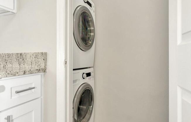 A stacked washer and dryer in a small laundry nook with light gray walls and a granite countertop nearby.