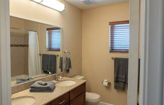 A clean, well-lit bathroom here at Dakota Ridge featuring a dual-sink vanity with granite countertops, dark wood cabinetry, a large wall mirror, neatly folded towels, a toilet, neutral wall tones, wood-style flooring, and a small window with blinds that brings in natural light.