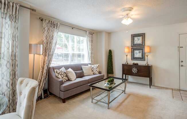 Staged living room with whit walls, large window with decorative curtains, carpeted floors, couch and coffee table, wall art, and entry table next to front door.