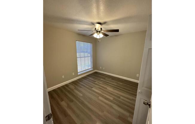 A spacious, empty room with light gray walls and new wooden flooring. A ceiling fan is centered in the room, which has a window with blinds allowing natural light to enter. The room is well-lit and has a simple, modern aesthetic.