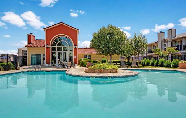 A resort-style swimming pool here at High Plains with clear blue water, surrounding lounge chairs, and a landscaped courtyard, complemented by a clubhouse building and bright skies that create a relaxing, inviting outdoor setting.