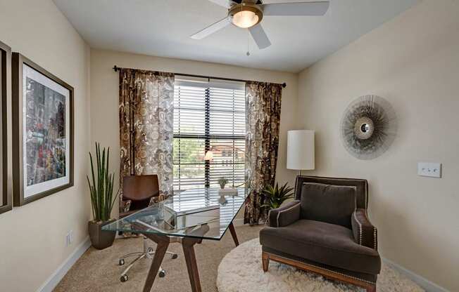 A bright home office here at Tuscany at Faudree with plush carpet, neutral walls, and a glass-top desk paired with a rolling chair. A comfortable accent chair sits on a soft area rug, while a ceiling fan and large window with blinds and patterned curtains bring in natural light and create a calm, functional workspace.