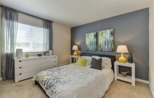 Bedroom with Large Window, White Comforter on Mattress, Lamps and Carpet at Monte Bello Apartments, Sacramento