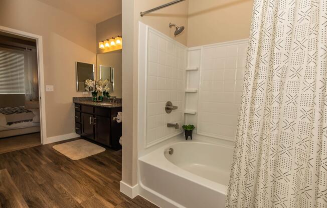 Modern bathroom featuring a shower-tub combination with a white shower curtain, a dark wood vanity with a large mirror, and stylish lighting. The floor is covered with wood-like vinyl, and there is a small plant on the edge of the tub. A doorway leads to a well-lit bedroom area in the background.