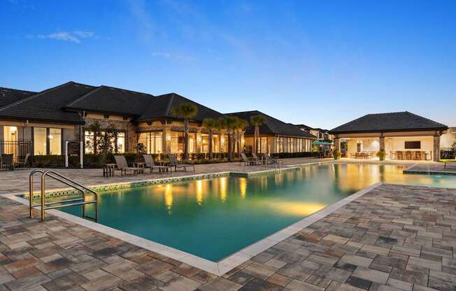 A large outdoor swimming pool surrounded by a stone patio and a building with a black roof.