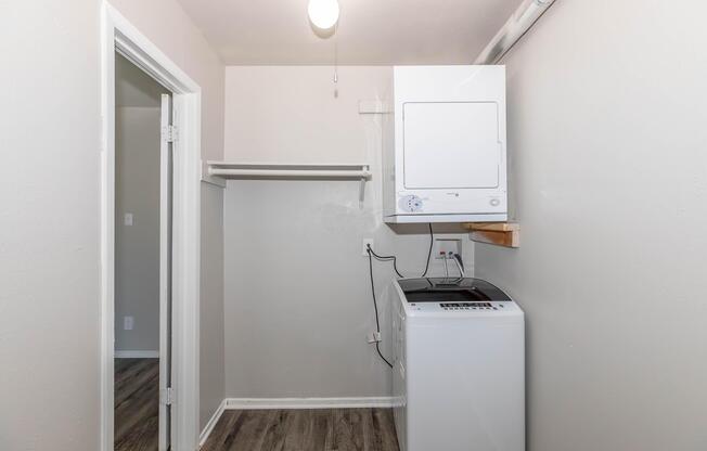 A laundry room featuring a stacked washer and dryer against a light gray wall. There is a white shelf mounted above the washer and dryer, and a doorway leading to another room on the left. The floor is finished with dark wood-like planks, and a ceiling light provides illumination.