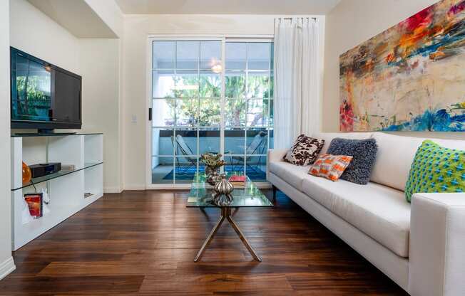 Living Room With Hardwood-Style Flooring, Sofa, Glass-Top Coffee Table, Media Cabinet with Glass Shelves, TV, And Sliding Glass Door Leading to Balcony. at Barrington Place, Rancho Cucamonga, CA