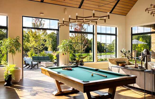 A pool table is in the middle of a room with a view of the trees outside.