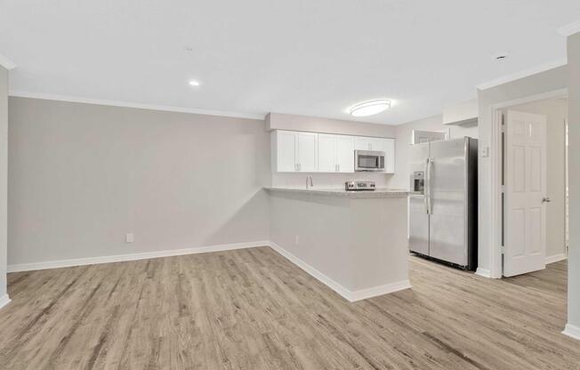 A spacious, modern kitchen and living area featuring light gray walls, wood-like flooring, and a kitchenette with white cabinets, a microwave, and stainless steel appliances. The area is well-lit with a circular ceiling light.
