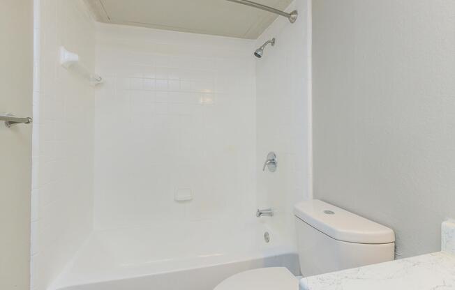 A clean and modern bathroom featuring a white tiled bathtub with a showerhead, a white toilet, and light gray walls. The space is well-lit and minimalistic, emphasizing a fresh and tidy appearance.