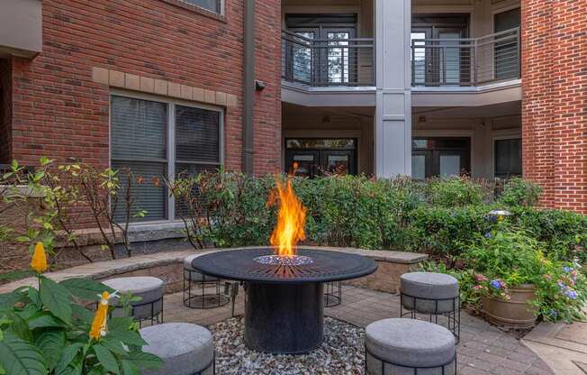 A fire pit sits in the middle of a courtyard surrounded by grey cushioned stools.