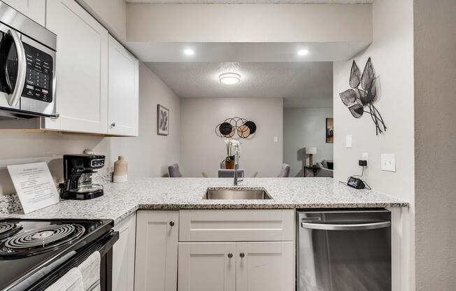 Modern kitchen featuring granite countertops, stainless steel appliances, and a coffee maker. There is a kitchen sink with a faucet, white cabinetry, and decorative wall art including leaf motifs. Soft lighting illuminates the space, creating a warm and inviting atmosphere.